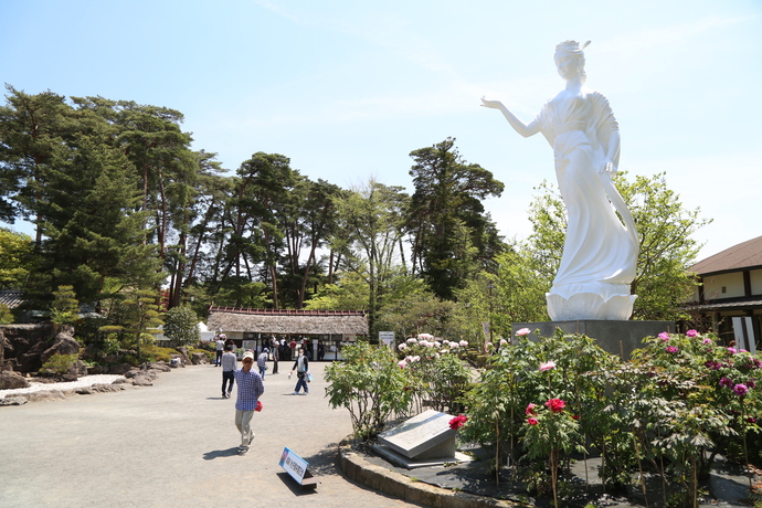 濃淡のピンク色の牡丹の円形の植栽の中央に真っ白な巨大な女性の像がそびえ立ち、複数の人々が歩く、緑に囲まれた広い道の奥には茅葺屋根の入り口が見える写真
