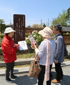 白い文字が刻まれた石碑が立った植栽の前で、男女の観光客に地図を見せながらガイドをしている赤い上着を着た女性の写真