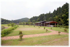 整備された芝生広場にレンガ造りの道があり、道の先に屋根付きのバーベキュー場、奥に森が広がっているフリーサイトの写真