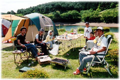 湖の前の芝生広場にテントを設営し、その前に置いたテーブルと椅子でくつろぐキャンプ客の写真