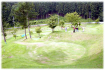 森を背景に、木々が点々と生えた緑の芝生のコースが広がるパークゴルフ場の写真