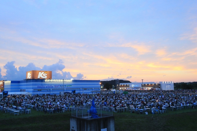 夕焼けに染まる淡いオレンジ色の雲が広がる空の下、大きな店舗ビルを背景にして、広大な広場を埋め尽くすほど大勢の人々がテーブルを囲んで集まり、賑やかにイベントを楽しんでいる夕暮れ時の風景の写真