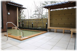 タイル敷きの清潔感のある露天風呂のスペースに、木枠で囲まれた手すり付きの湯船、休憩用のベンチが設置されており、周囲を竹垣と木々に囲まれた露天風呂の写真