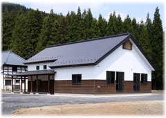 森を背景に建つ、濃いグレーの屋根で、壁の上部が白壁、下部が茶色の木目調で構成された和風建築のふるさと体験館の外観写真