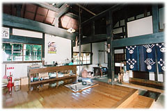 高い天井の空間に、囲炉裏が設置された板の間、かまどのある台所へ続く土間があり、奥には青い暖簾が掛かった入り口が見える写真