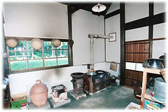 煙突の付いた古いかまどや火鉢、大きな水瓶などが置かれ、窓の外には緑豊かな景色が広がる土間の炊事場の写真