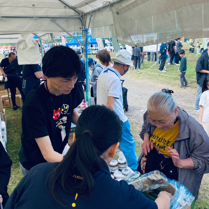 キュウリなどを販売している白いテントの下で、女性の買い物客の対応をしている、黒いTシャツの男性と紺色のポロシャツの女性の写真