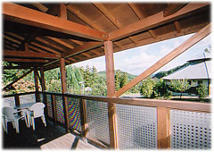 白いテーブルセットが置かれたバルコニーの屋根と手すりの間から、木々や遠くの山々の開放的な景色が見える写真