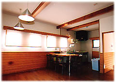 木目調の腰壁が付いた白い壁の部屋で、フローリングの床に黒いキッチンが置かれ、天井からペンダントライトが下がったダイニング・キッチンの写真