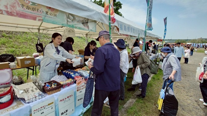 屋外に並んだ、キャラクターのグッズなどを販売している白いテントに複数の客が集まり、女性の販売員が男性に商品を手渡している物販の様子の写真