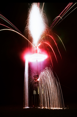 暗闇の中に設置された仕掛けから、激しく火花を吹き上げる花火や回転する花火、滝のように地面へ流れ落ちる花火、上空へ弧を描いて広がる赤や白の花火が夜空を彩る、ダイナミックな仕掛け花火の写真