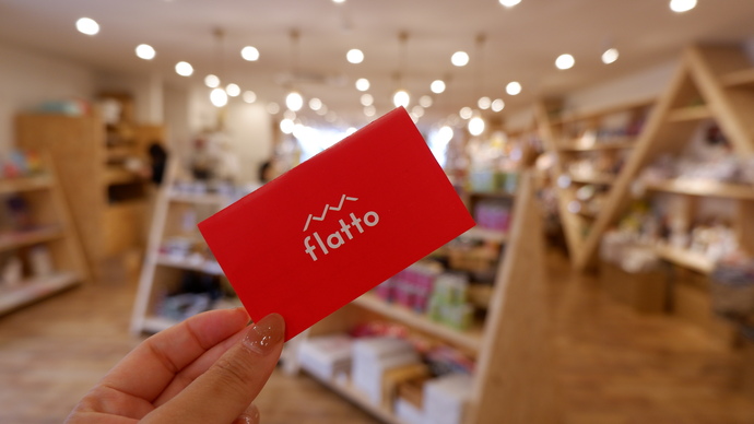 雑貨店のような場所を背景に、女性の手が持ち上げた赤いflattoポイントカードにフォーカスをあてた写真