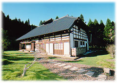 豊かな緑の木々を背景に、黒い瓦屋根で、白い壁に木が格子状にあしらわれた日本家屋が建ち、手前に広がる芝生の庭に石畳の通路が設けられた本館の外観写真