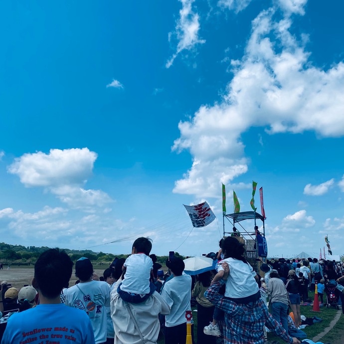 青空の下、広場に集まった大勢の観客の中、子どもたちが大人の肩車に乗って「葵陽」の凧があがる様子を見物し、凧の後ろには2人の人物が乗った小さな櫓がある、まつり会場の写真