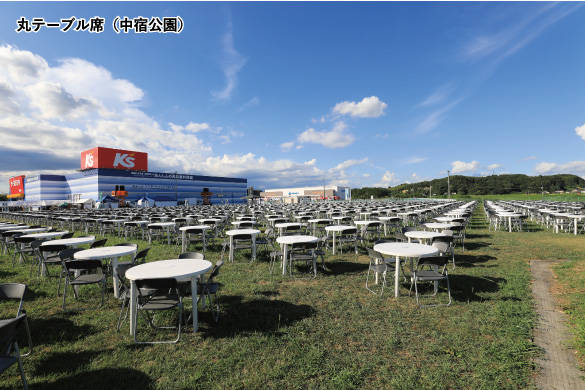 澄み渡る青空の下、ケーズデンキの店舗が隣接する広大な芝生の中宿公園に、白い丸テーブルと黒い椅子が整然と並べられている、丸テーブル席（中宿公園）の写真