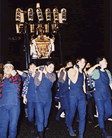 夜の暗闇の中、揃いの青い腹掛けや股引を身にまとった担ぎ手たちが、上部に提灯を掲げた黄金色に輝く立派な神輿を肩に担いで練り歩いている写真