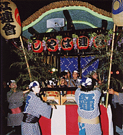 夜の闇の中、揃いの法被を粋に着こなした人々が、色とりどりの提灯や藤の花のような装飾で彩られた華やかな山車を囲み、伝統的な祭りの囃子を奏でながら賑やかに練り歩いている祭りの風景の写真