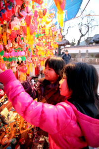 屋台に色鮮やかな飾り物がぎっしりと並ぶ通りで、2人の子どもが興味深そうに手を伸ばして好みの品を探している様子の写真