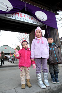門前で三人の子どもが冬服を着て並び、境内のにぎわいを背景に元気に立つ様子を写した写真