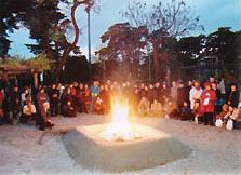 木々に囲まれた広場で、四角い盛り土の中央で燃える炎を大勢の人が囲んで見つめている牡丹焚火の様子の写真