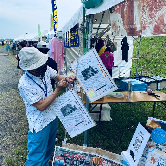 まつり会場に並んだ白いテントの下で、白い帽子と白地に水玉柄のシャツを着た男性が、ラミネートされた新聞記事の切り抜きをテントの柱に下げる作業をしている様子の写真
