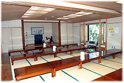 広々とした和室にたくさんの座卓が整然と並べられており、奥の床の間にはテレビが置かれている大広間の写真