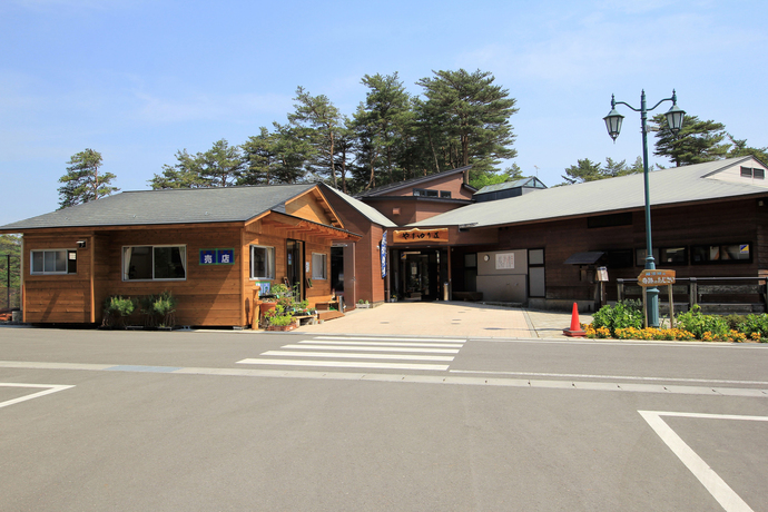 松林を背景に建つ平屋の木造で、手前の駐車場から入り口へ続く舗装された通路や、横断歩道、街灯、手入れされた花壇が並ぶ、やまゆり荘の外観写真