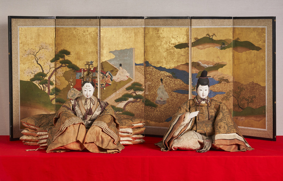 金屏風を背景に、赤い毛氈の上に豪華な衣装を着た二体の古今雛人形が並び、左側は冠を付けた人形、右側は烏帽子をかぶり巻物を持つ人形で、屏風には山水や松、梅が描かれた雅な装飾が施されている写真