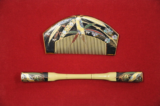 赤い布の上に黒と金を基調とした蒔絵の櫛と笄が並び、櫛には草花や流水を描いた華やかな装飾が施され、笄にも同様の模様が両端に描かれた伝統的な和装用髪飾りの写真