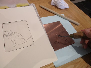 木製のテーブル上で銅版に猫の線画を彫る作業が行われており、右側には彫刻刀を持つ手、左側には同じ猫の図柄が刷られた紙が置かれ、背景には白い棒状の道具や布が見える版画制作の様子の写真
