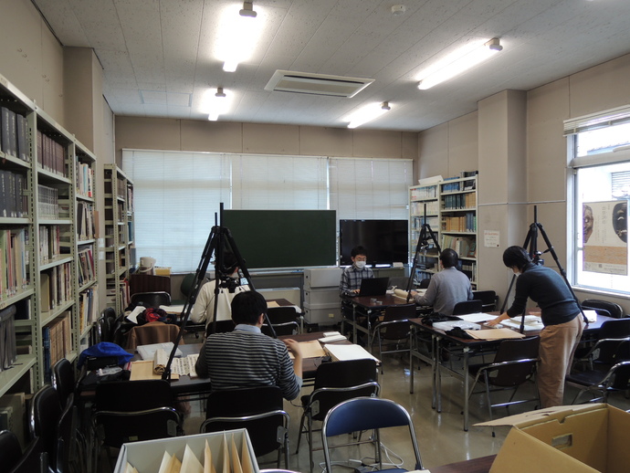壁際に多くの文献が並んだ棚が複数置かれ、黒板やモニターが奥に配置された部屋に複数の長机やいすが並び、5人の人物がそれぞれのテーブルに資料を広げ作業をしている、古文書調査の様子の写真