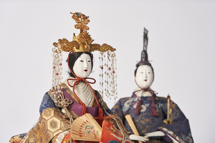 豪華な衣装を着た二体の古今雛人形が並び、手前は金色の冠に垂飾を付け、扇を持ち紫や赤を基調とした刺繍入りの衣装を着用し、奥は烏帽子をかぶり巻物を持ち青系の文様入り衣装をまとった雅な雰囲気の写真