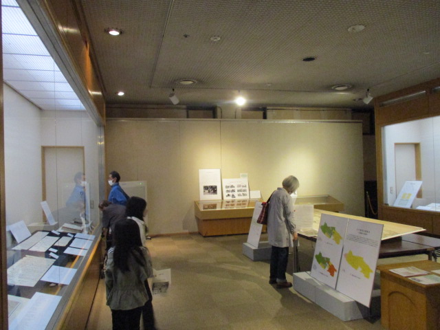 部屋の奥や中央に置かれた展示台や左右の展示ケースに資料が展示され、複数の来場者が見学をしているテーマ展の様子の写真