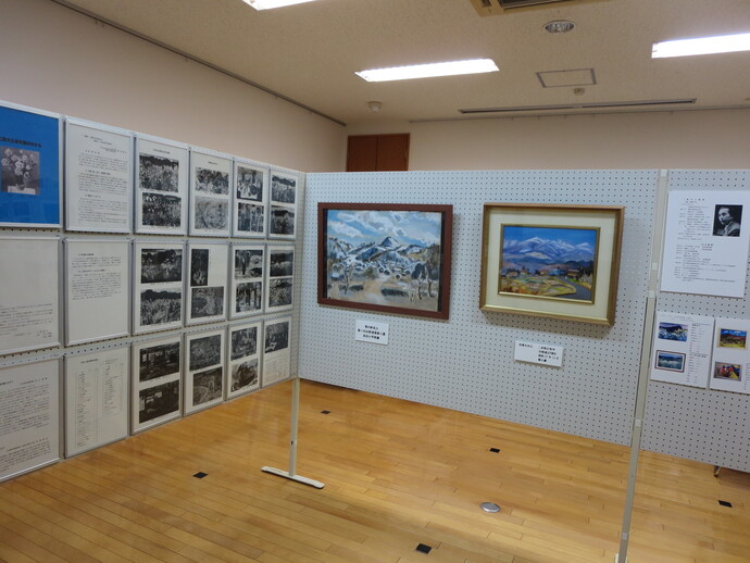 明るい屋内の展示会場で、木目調の床に設置された白いパーテーションに、風景を描いた2枚の額装絵画や、多くの資料写真、説明テキストが整然と並べて掲示されている、展示会の設営風景を捉えた写真