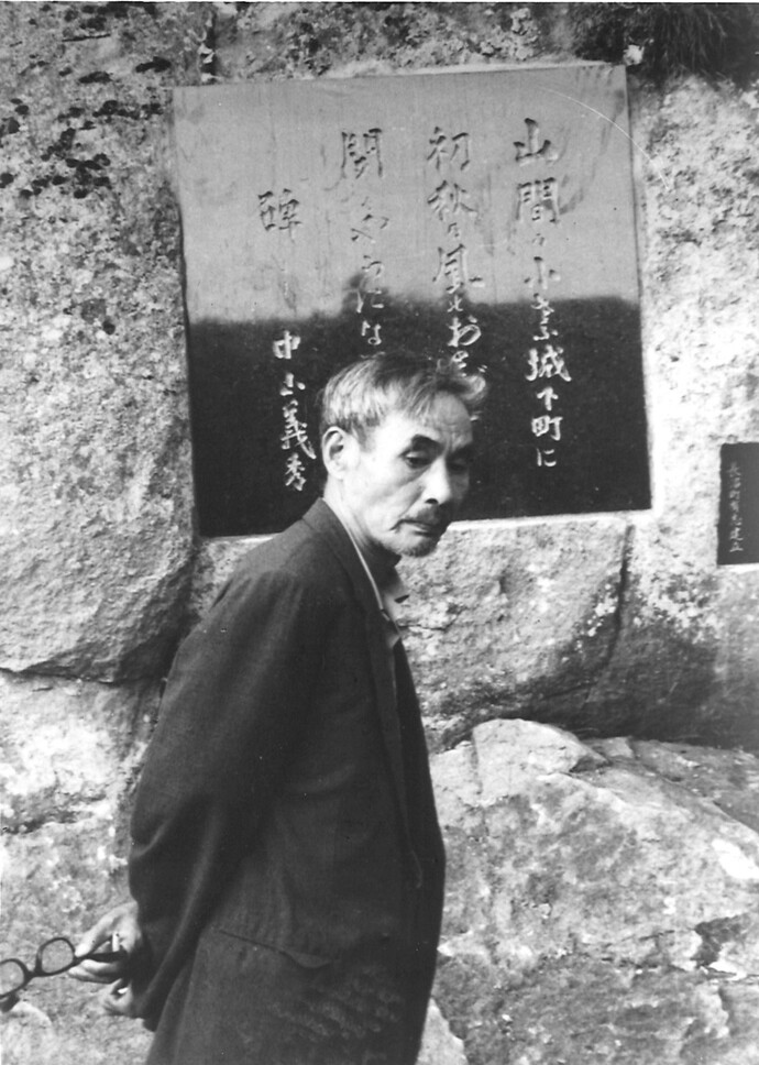 背広姿の中山義秀が、ゴツゴツとした岩肌に埋め込まれた、達筆な日本語の詩や人名が刻まれている大きな石碑の前に立ち、物憂げな表情で傍らを見つめている様子を捉えたモノクロの写真