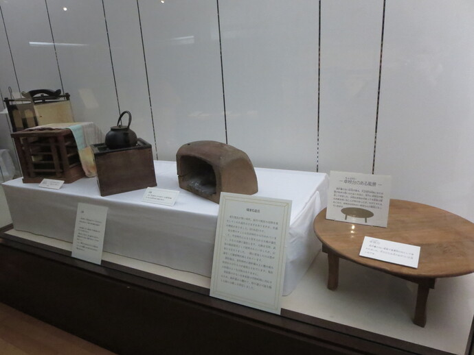白布が敷かれた展示台の上に鉄瓶が載った火鉢や土製の竈、さらにその横には木製のちゃぶ台が置かれ、かつての家庭の台所や居間の風景を伝える解説パネルと共に紹介されている生活用具の展示風景の写真