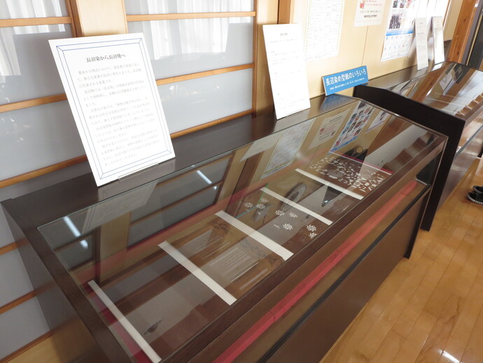 黒縁の横長なガラス展示ケースの中に、植物や幾何学模様が細やかに彫り抜かれた数枚の茶褐色の型紙が並べられ、ケースの上には「長沼染め型紙のいろいろ」という案内や解説パネルが添えられている展示風景の写真