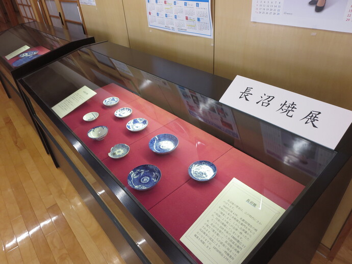 黒縁のガラス展示ケースの中に藍色の絵付けが施された様々な大きさの小皿が赤い布の上に並べられ、「長沼焼展」と記された案内板や歴史を解説するパネルと共に紹介されている展示風景の写真