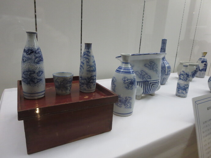 木製の台の上に置かれた藍色の風景模様が描かれた徳利とお猪口に加え、周囲にも様々な形状の染付磁器が白布の上に整然と並べられ、壁際の解説パネルと共に紹介されている陶磁器の展示風景の写真