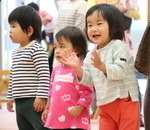 室内で3人の幼児が並んで立っており、手前の子どもが笑みを浮かべて両手をたたく様子の写真