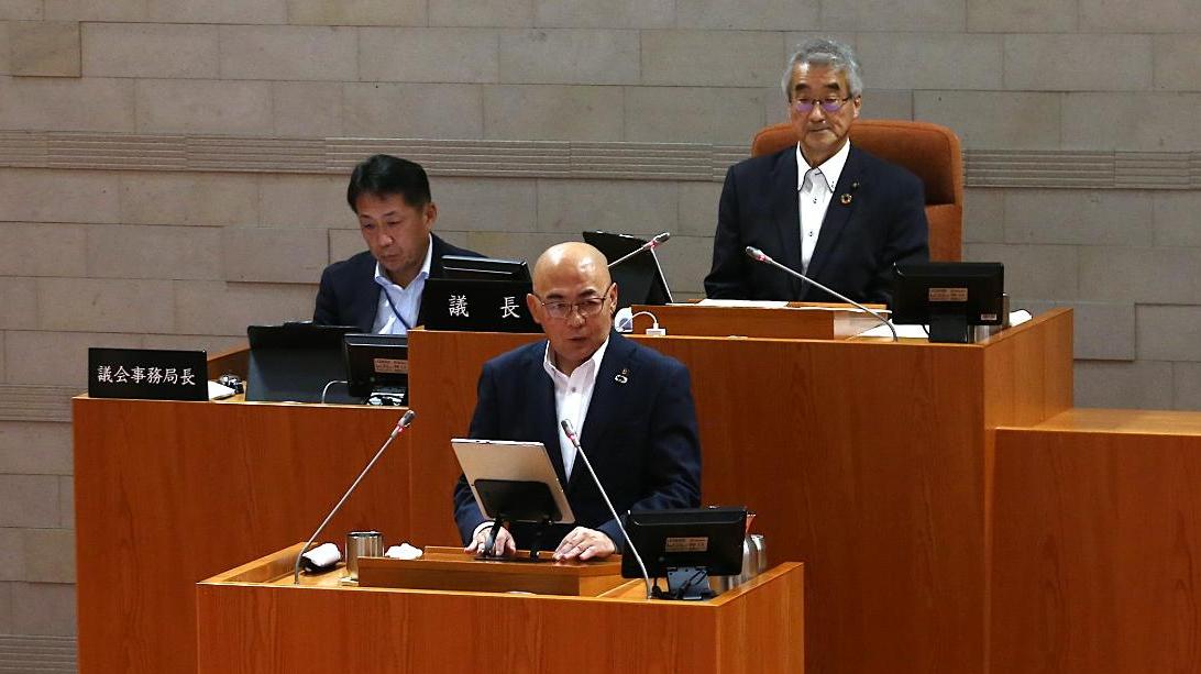 演壇に立ち答弁を行うスーツ姿の男性と、その後方の席に座る議長などと思われる二人の男性が議場に並び、厳かな雰囲気の中で会議が進行している様子を捉えた写真