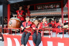 祭りの屋台風の舞台で赤い衣装を着た子どもたちが並び、複数の和太鼓を前にバチを持って演奏準備をしている様子が写された写真