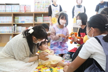 マットの上に座った乳児を母親が優しく支え、その周りをマスク姿の学生たちがぬいぐるみや玩具を使ってあやしながら交流している、育児支援や実習の様子を捉えた写真