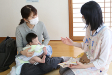 畳の部屋で、赤ちゃんを抱いたマスク姿の母親が、エプロンを着用した支援員の女性と向き合い、育児に関する相談やアドバイスを熱心に受けている様子の写真