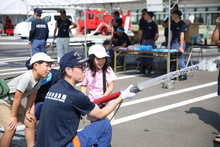 紺色の制服を着た消防団員が膝をつきながら放水ホースを支え、その隣で子供たちが放水の様子を間近で見守っている、地域の防災訓練での体験学習シーンを捉えた写真