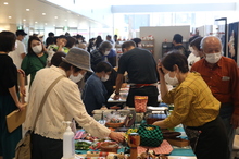 明るく広々とした屋内の展示会場で、マスクを着用した大勢の来場者がテーブルに並べられた色とりどりの雑貨や工芸品を熱心に手に取り、出店者と交流しながら買い物を楽しんでいる、マーケットの様子を捉えた写真