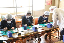 衛生キャップとマスクを着用した制服姿の生徒たちが、指導員に見守られながら机に並び、はんだごてを手に精密な電子基板の組み立て作業に真剣な表情で取り組んでいる様子の写真