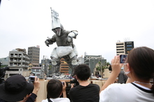 曇り空のミニチュアの市街地を背景に、頭が尖った怪獣のオブジェが設置されており、手前にいる観客たちがスマートフォンやカメラを向けてその姿を熱心に撮影している、屋外イベントの様子を捉えた写真