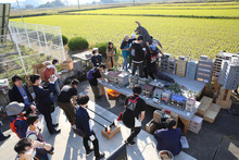 広大な田園風景を背景に、屋外に設置された精巧なミニチュアの街並みのセットで、スタッフたちが怪獣のスーツを調整したり撮影準備を進めたりしている様子を、見学者たちが周囲から興味深く見守っている特撮映画の撮影現場を捉えた写真