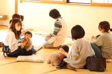 陽の光が差し込む畳敷きの明るい室内で、数組の母親と乳幼児たちが思い思いの場所に座り、スタッフに見守られながら和やかに過ごしたり育児相談をしたりしている、地域の子育て支援施設の活動風景を捉えた写真